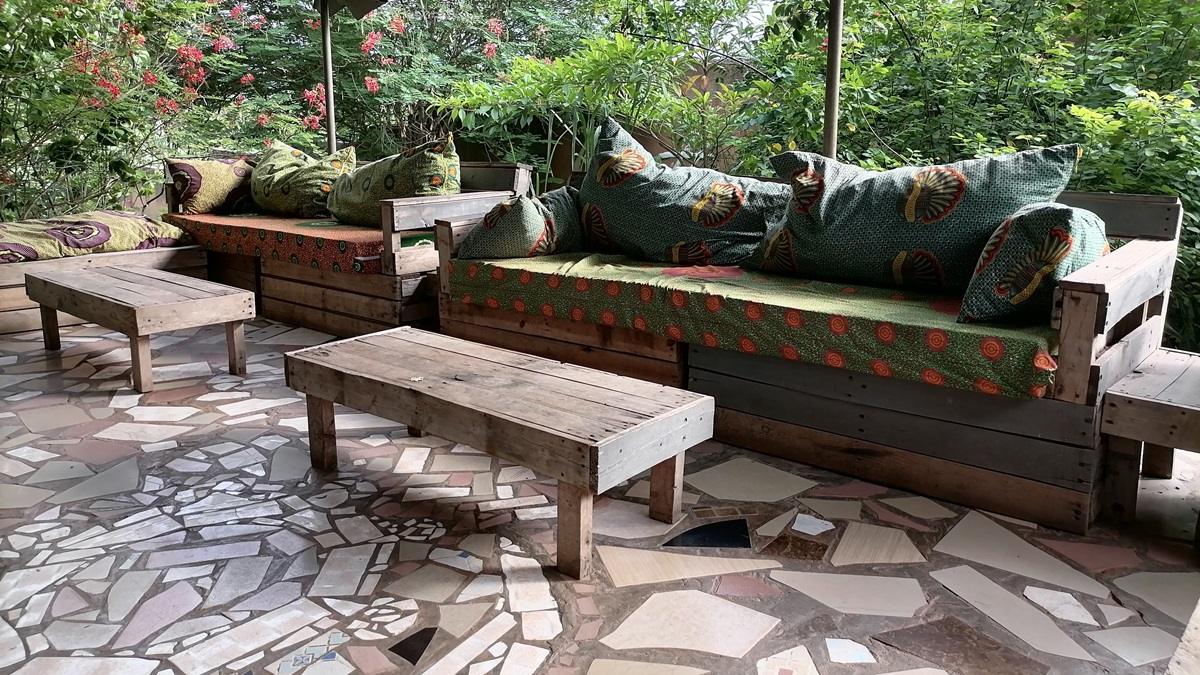 A pallet sofa with a colorful African print cushion and pillows on a porch surrounded by plants.